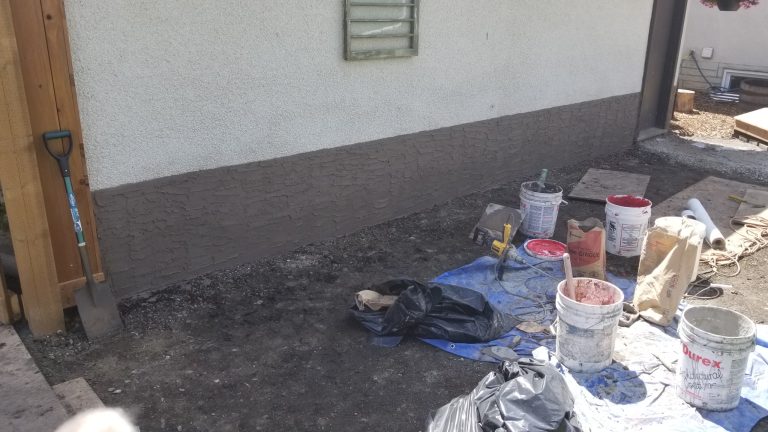 Parging repair process in Edmonton, showing freshly applied brown parge on foundation wall, with construction materials and tools including buckets, trowels, and tarps nearby.