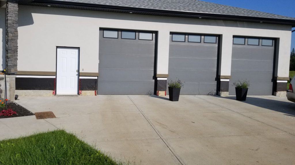 Modern garage with three gray doors, a white side door, and planters, showcasing stucco exterior and driveway, relevant to stucco repair services.