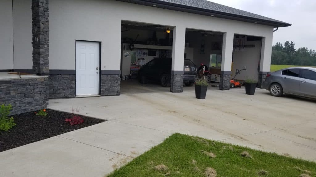 Modern garage with stone accents, featuring two open bays, parked vehicles, and a white door, surrounded by landscaped grass and flower beds, illustrating a residential property suitable for stucco repair services.