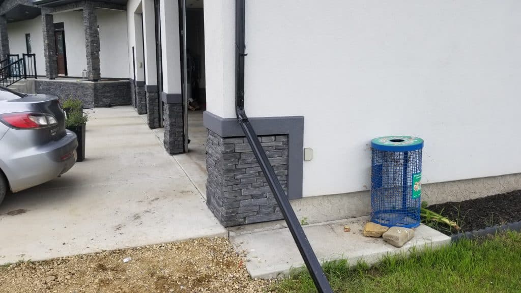 Exterior view of a residential building featuring stucco and stone accents, with a gray car parked nearby, a drainage pipe leading to a wall, and a blue trash can positioned on the ground.