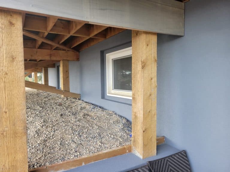 Acrylic parging installation showcasing a gray exterior wall with a window, wooden support beams, and gravel ground, emphasizing the aesthetic appeal and protective qualities of acrylic finishes.