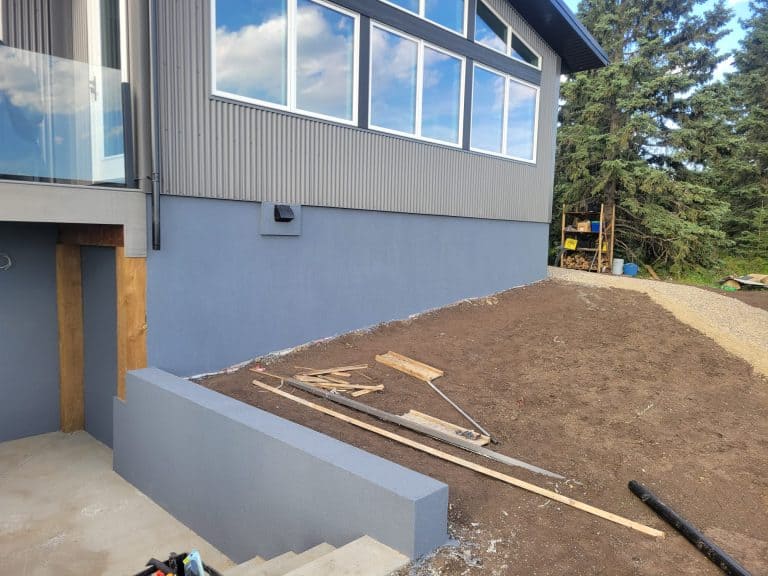 Acrylic parging installation on a modern home exterior, showcasing freshly applied gray coating, adjacent construction materials, and a landscaped area.
