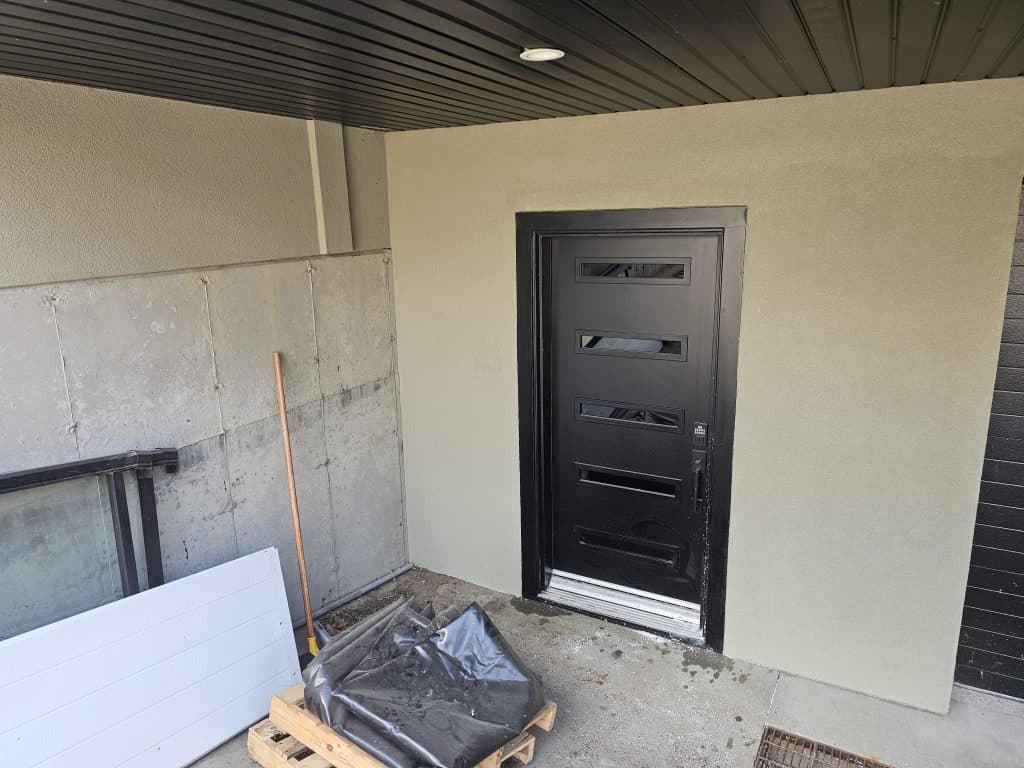 Black modern door with horizontal glass panels, adjacent to newly applied EIFS stucco finish, construction materials including a wooden pallet, plastic sheeting, and a broom in a residential entryway setting.